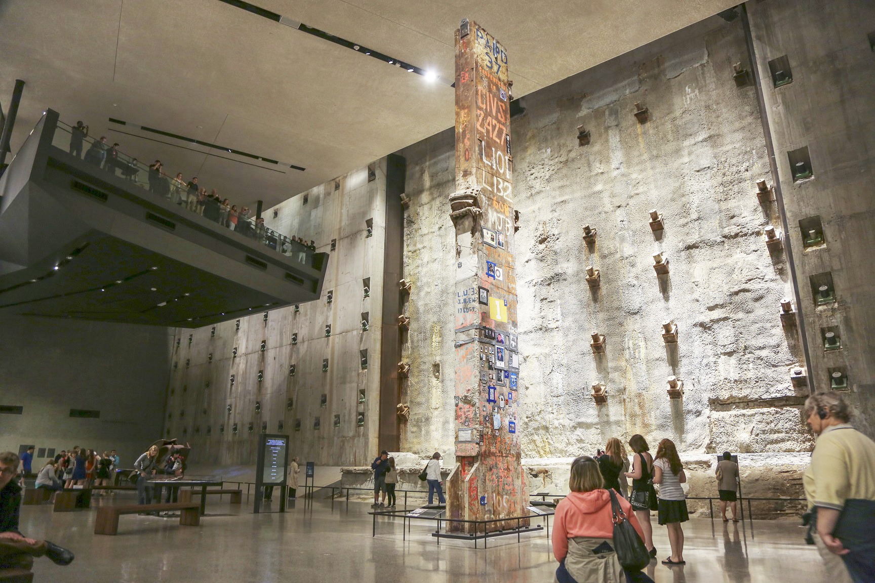 The Last Column stands over Foundation Hall as visitors walk by it. The illuminated slurry wall stands in the background.