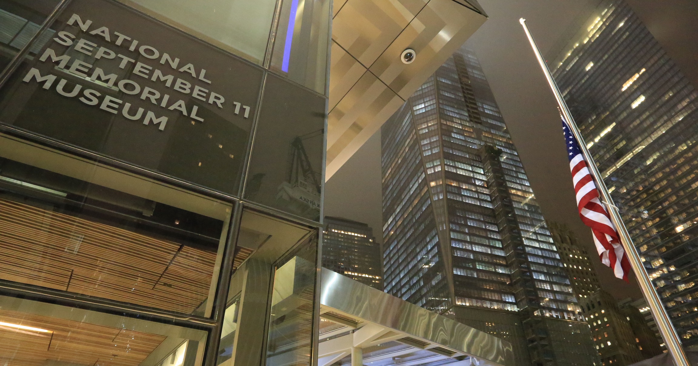 The entrance to the 9/11 Memorial & Museum is seen illuminated at night. One World Trade Center and an American flag at half-staff stand off to the right.