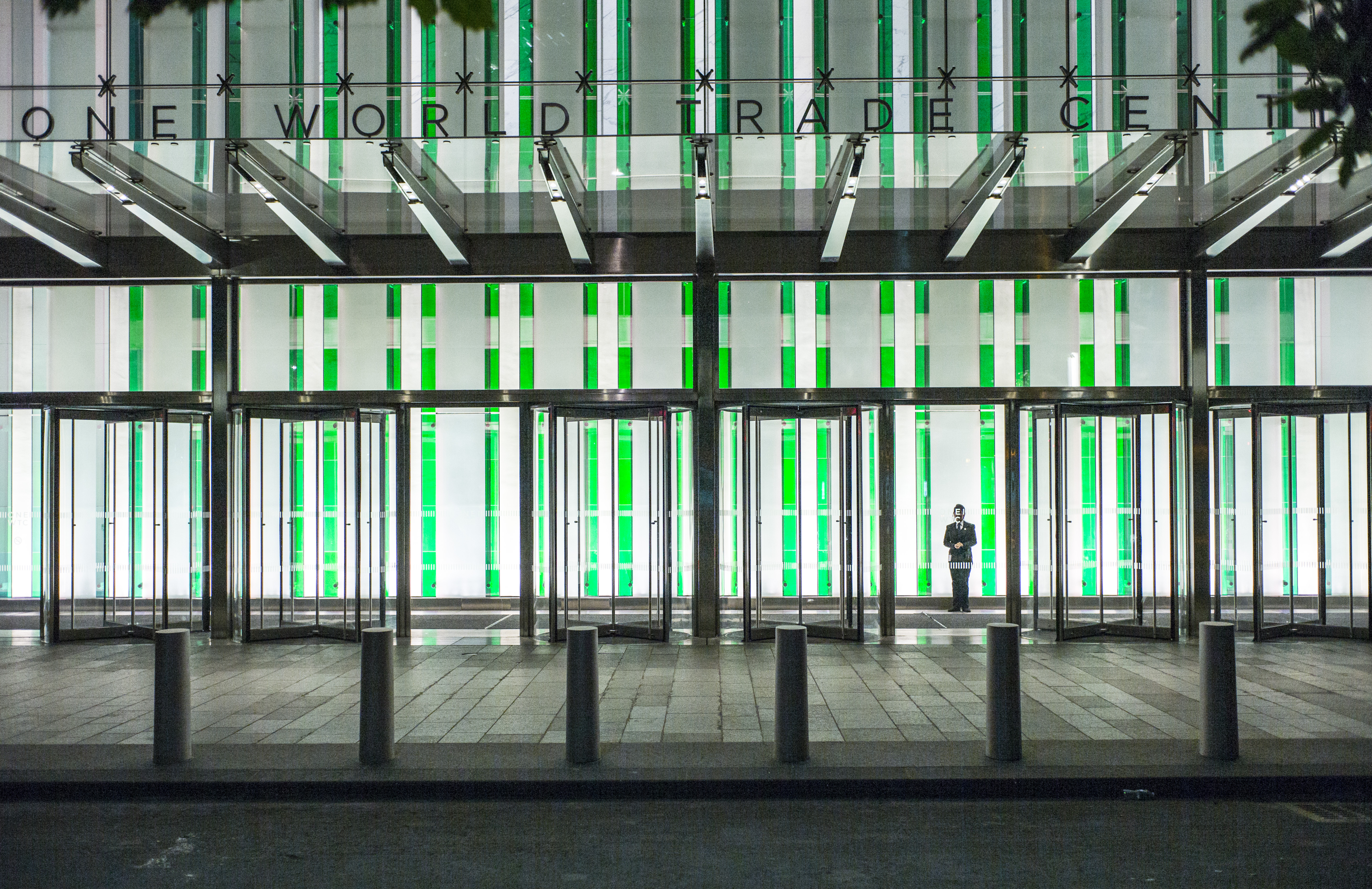 The entrance to One World Trade Center is illuminated at night. A man in a suit and tie stands behind the glass entrance doors.