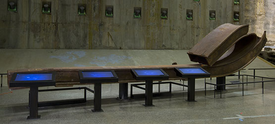 A large rusty beam from the Twin Towers is displayed beside four signing screens in Foundation Hall. The concrete slurry wall is visible in the background.