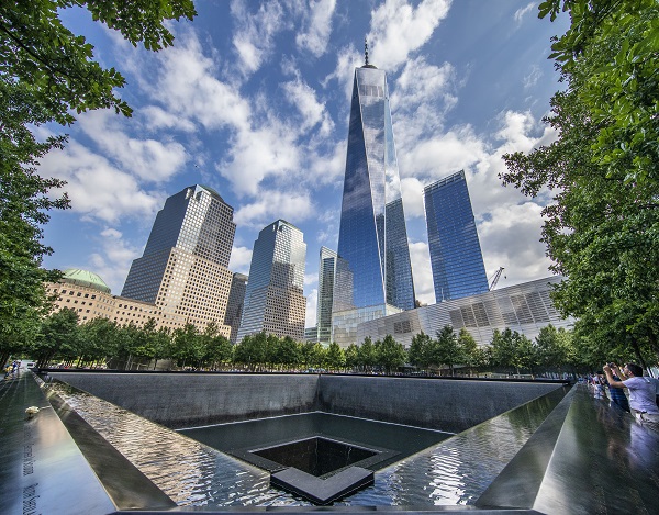 One World Trade Center and surrounding skyscrapers tower over Memorial Plaza. Clouds and a blue sky reflect on One World Trade Center and on the water of the south pool.