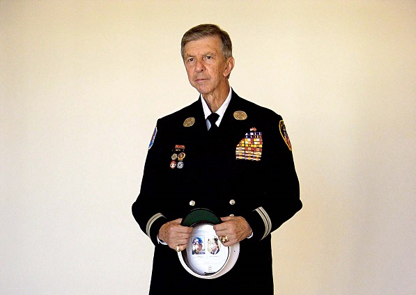 Retired FDNY Captain John Vigiano Sr. poses in a formal outfit. He holds a hat that features two images of his sons, who died on 9/11.