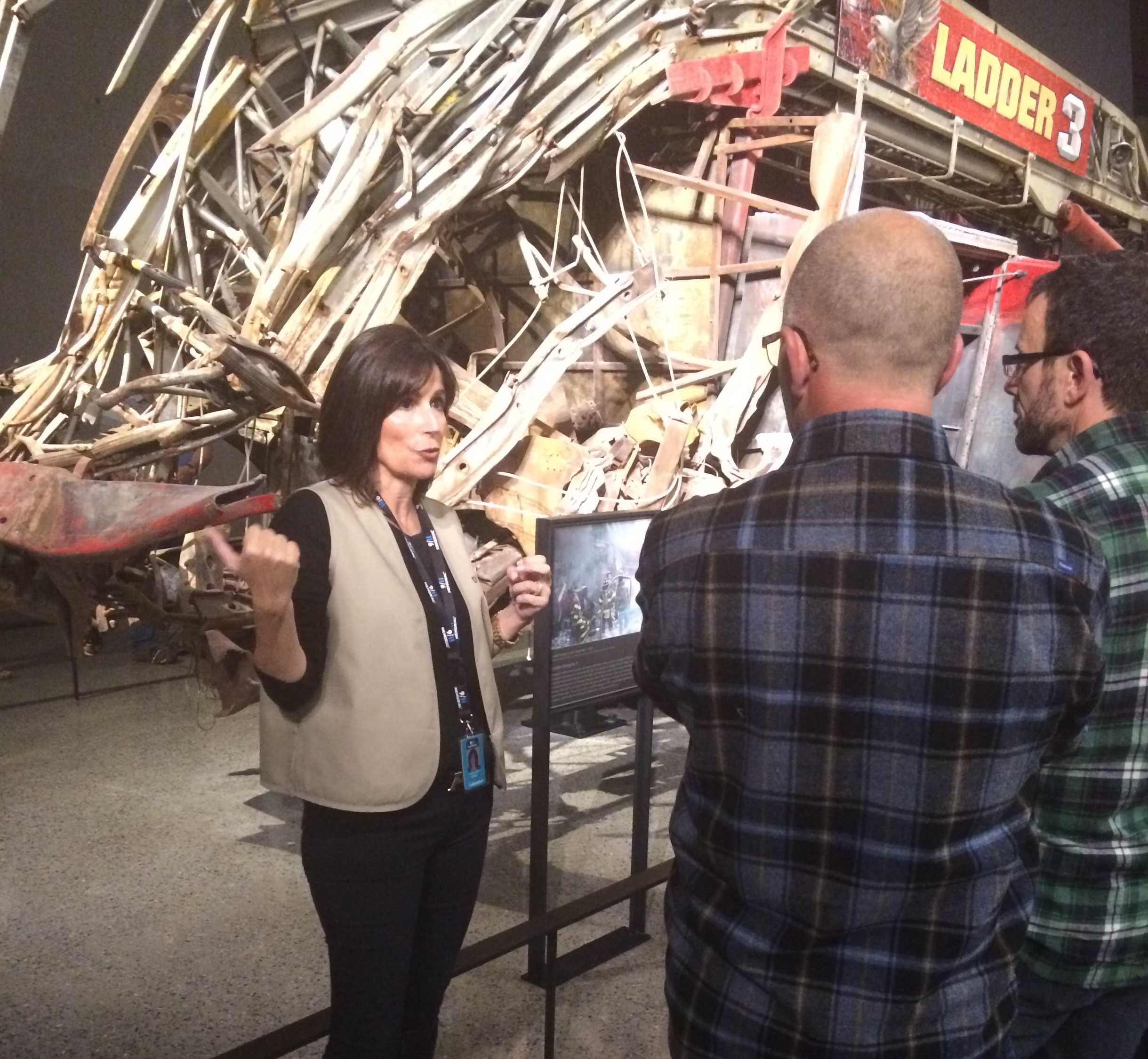 Maria Jaffe, a participant in the 9/11 Memorial Museum’s docent program, gives a tour to two men visiting the Museum. A heavily damaged fire engine is on display behind her.