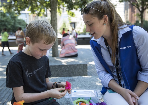 A young boy makes origami as a young woman in a volunteer vest watches on. The two are sitting on a bench at the Memorial as part of the Stories & Art program.