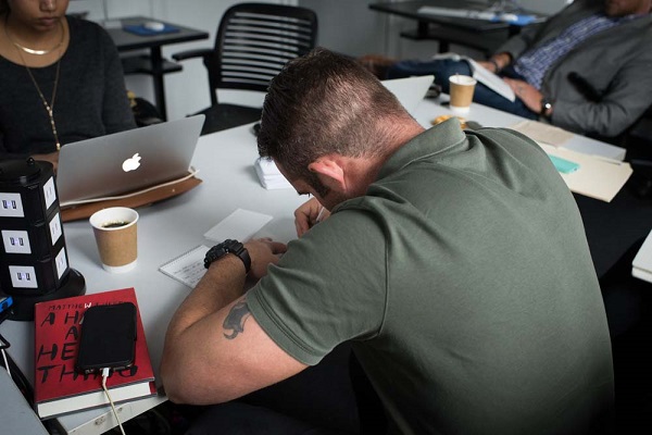 Derek McElroy, a War Horse writing fellow, writes in his notebook while seated at a table. A woman across from him is on her Apple laptop.