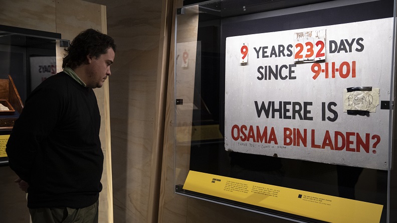 A visitor peers into a glass wall case containing a homemade sign that reads, "9 years 232 days since 9-11-01 Where Is Osama bin Laden?" in the museum exhibition "Revealed: The Hunt for Bin Laden."