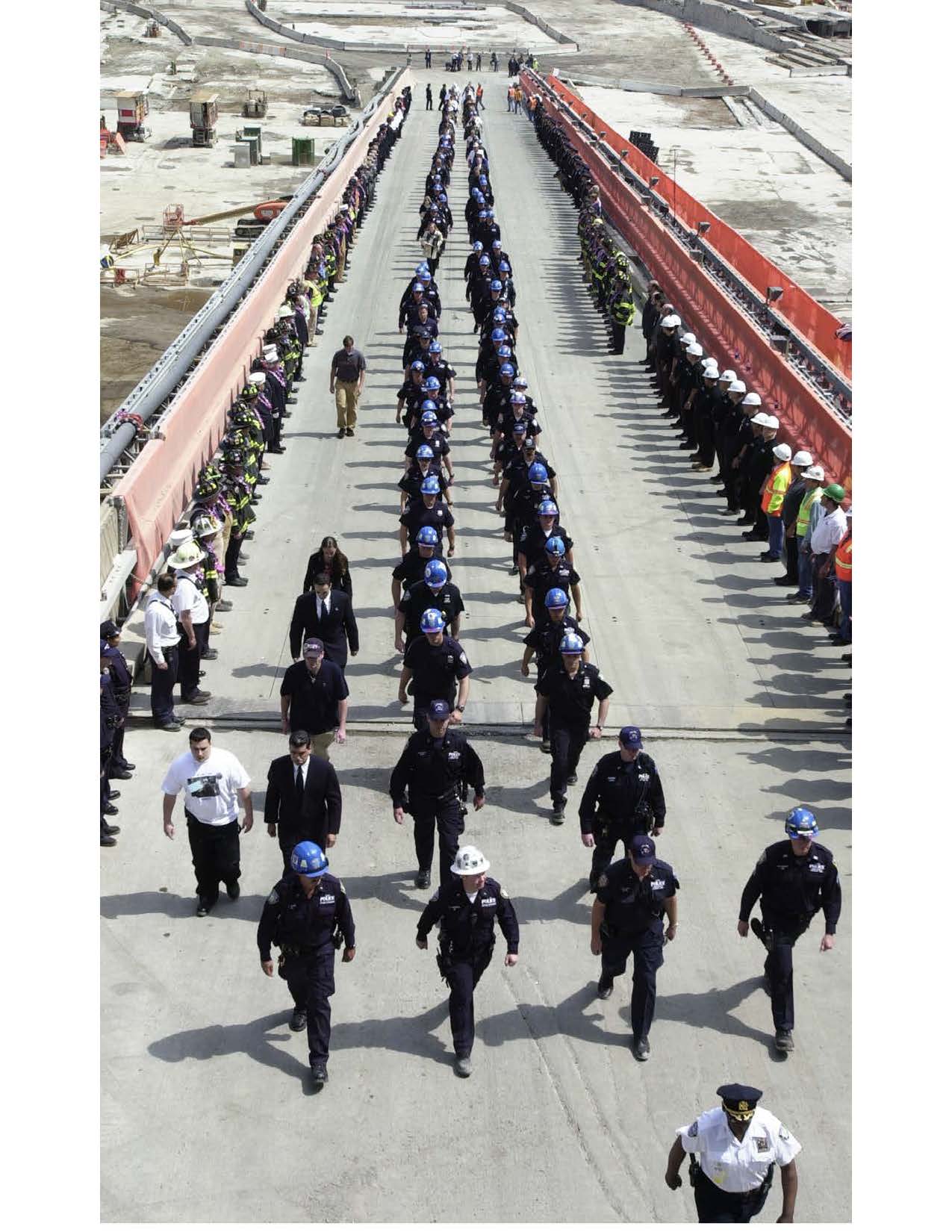 Uniformed first responders wearing hard hats walk in two neat rows away from the Ground Zero site.