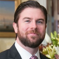Bearded man with light brown hair in a white shirt, navy blue jacket and maroon tie