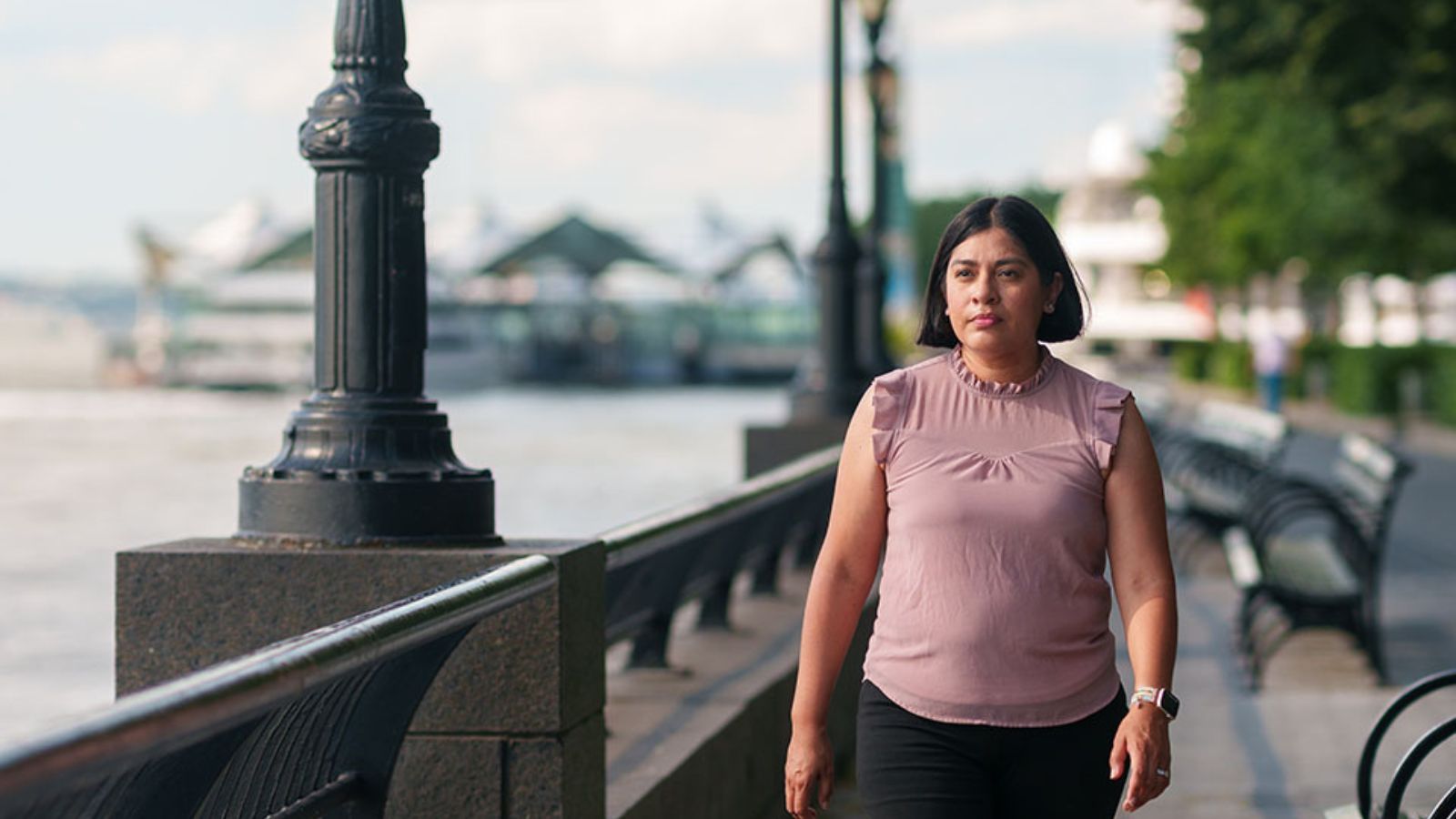 A woman with chin-length dark hair, wearing a pink sleeveless top and black jeans, walks along the water with benches to her left