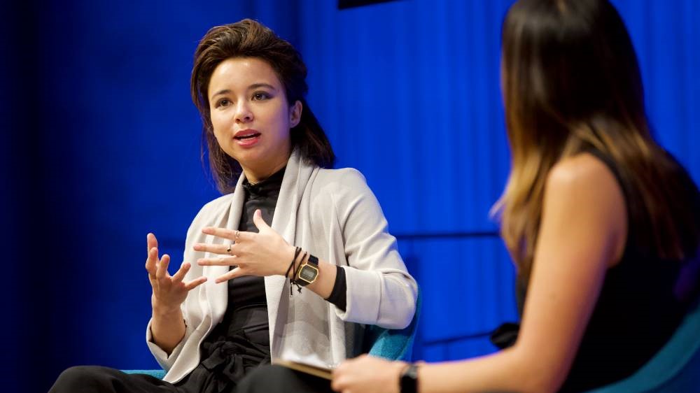 VICE correspondent Isobel Yeung gestures as she speaks onstage next to a woman hosting a public program at the Museum Auditorium. A curtain behind her is lit up blue.