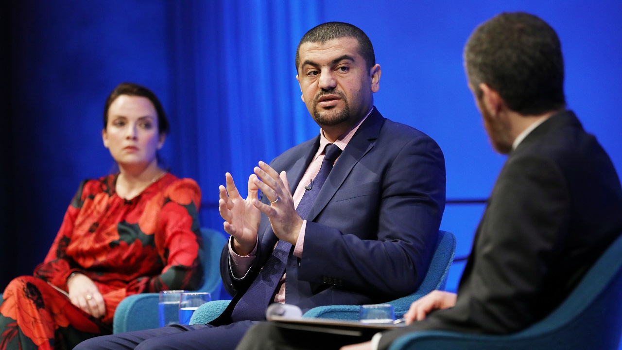 Journalist Hassan Hassan gestures with both hands as he speaks to the 9/11 Memorial & Museum’s Noah Rauch, who is moderating a public program at the Museum Auditorium. Rauch is in the foreground and out of focus. In the distance to Hassan’s right is American Enterprise Institute scholar Karen E. Young, who is listening as Hassan speaks.