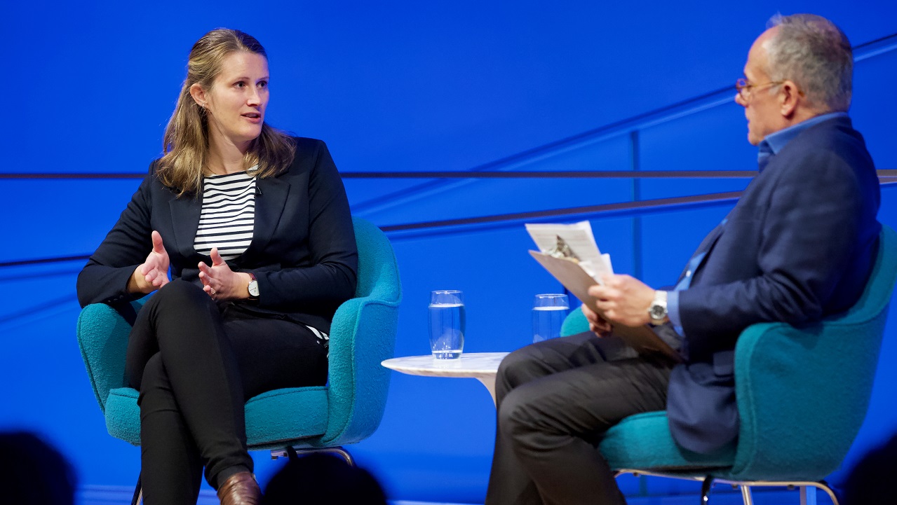 A female speaker gestures with her hands on a blue-lit auditorium stage while the male moderator looks on.