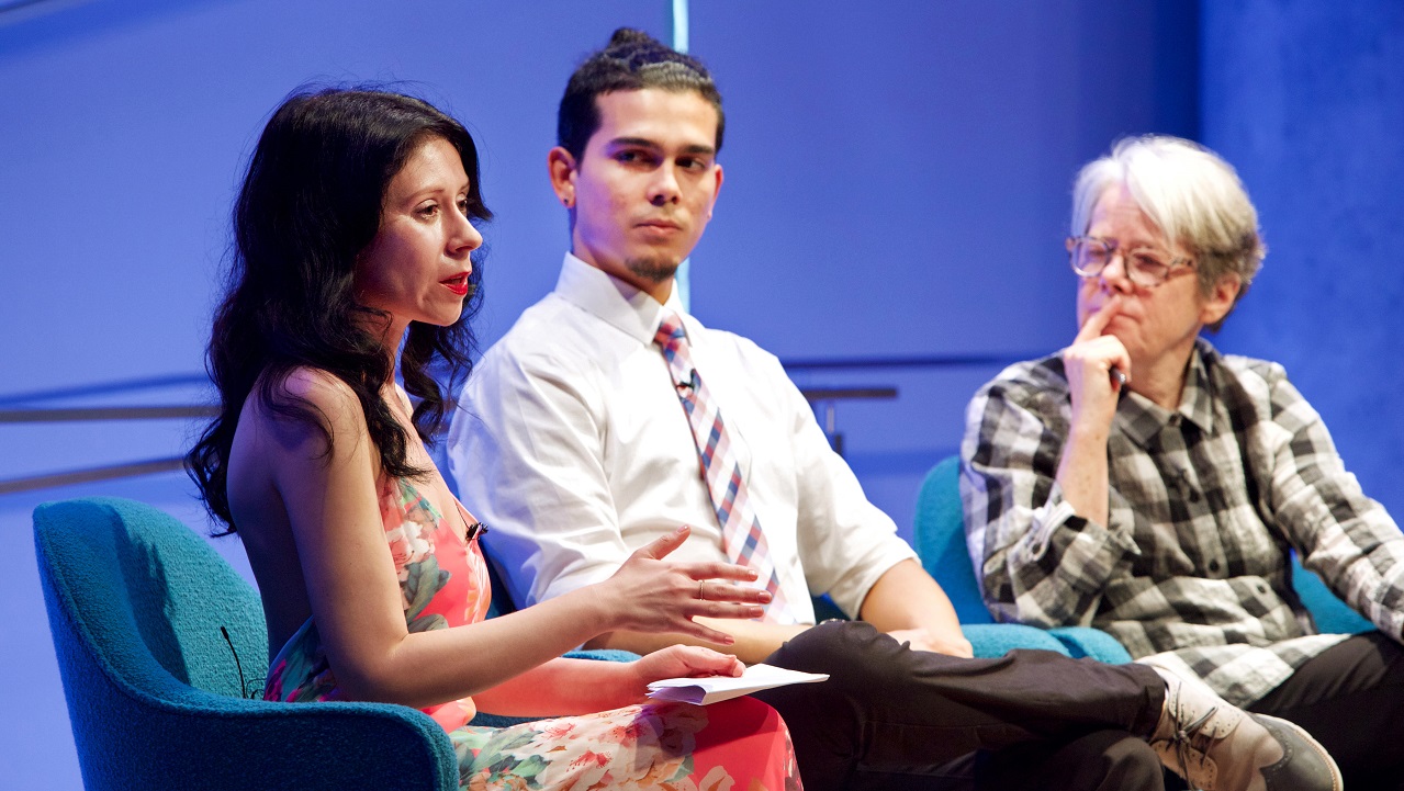 Four people sit on a blue-lit auditorium stage during a public program.