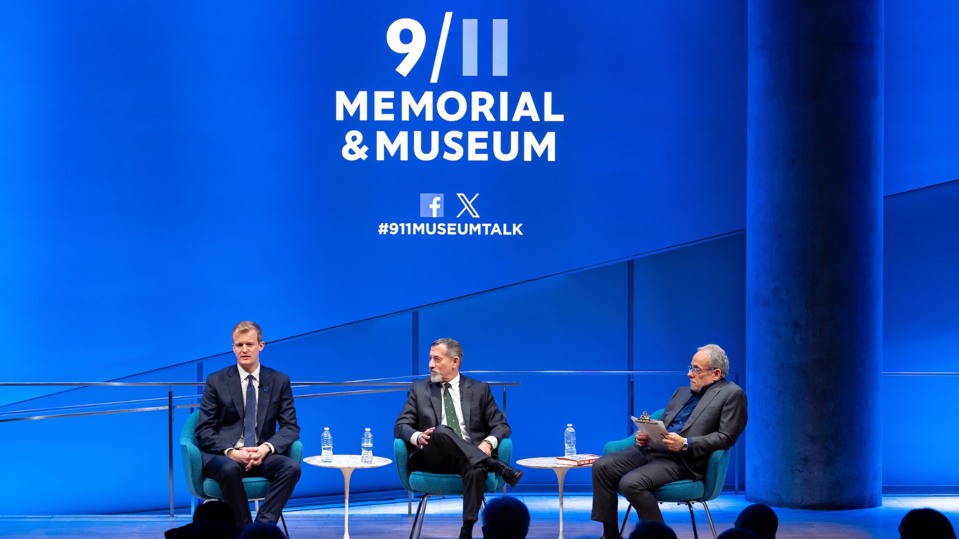 Three men on stage, all wearing dark suits, against a blue background displaying the 9/11 Memorial and Museum logo.
