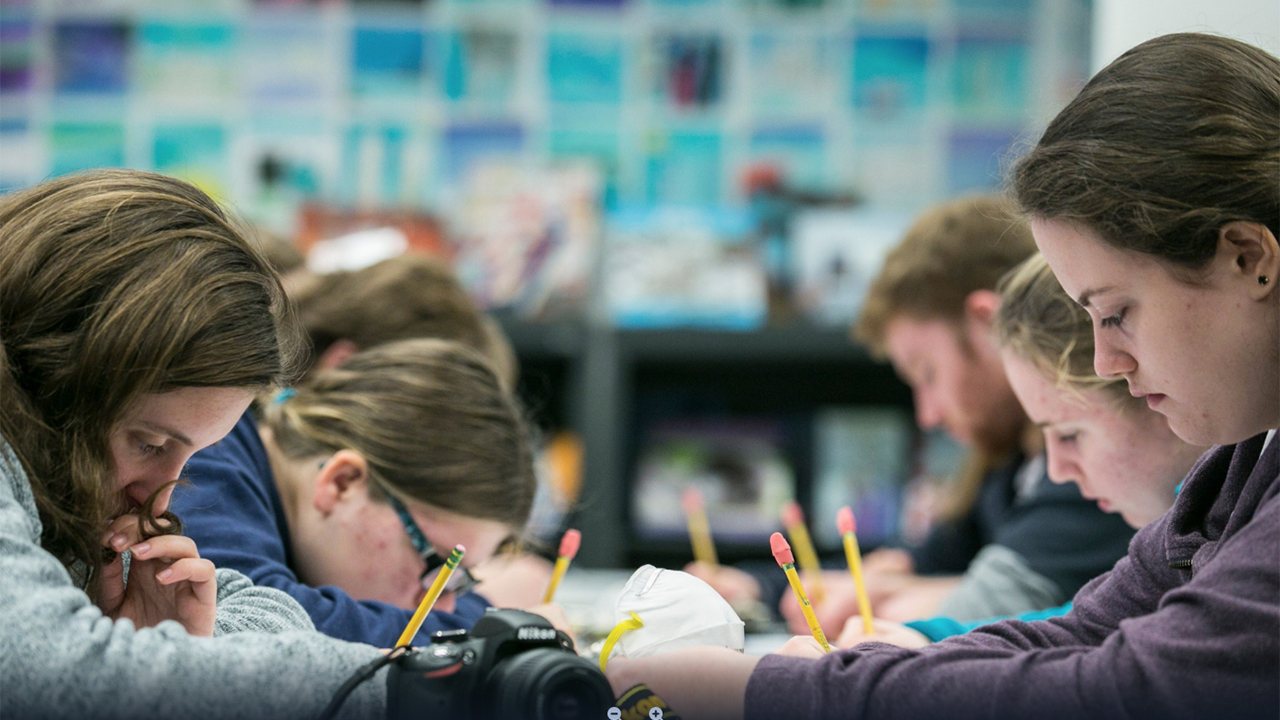 A group of students sit around a table, holding pencils, and peering down as they work. They are deeply engaged in a learning activity.