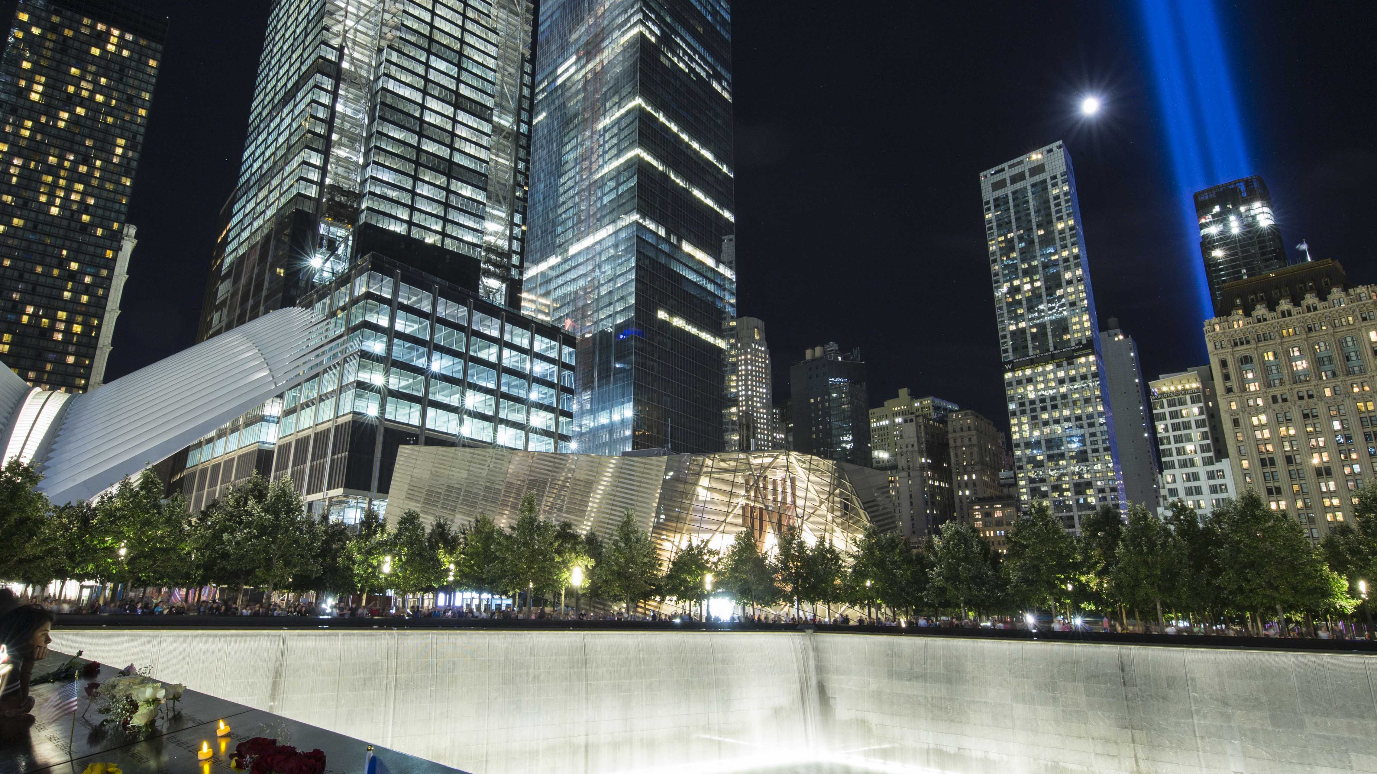 The twin beams of the Tribute in Light shine beside the moon over the buildings of lower Manhattan. In the foreground is the North Tower reflecting pool of Memorial Plaza.