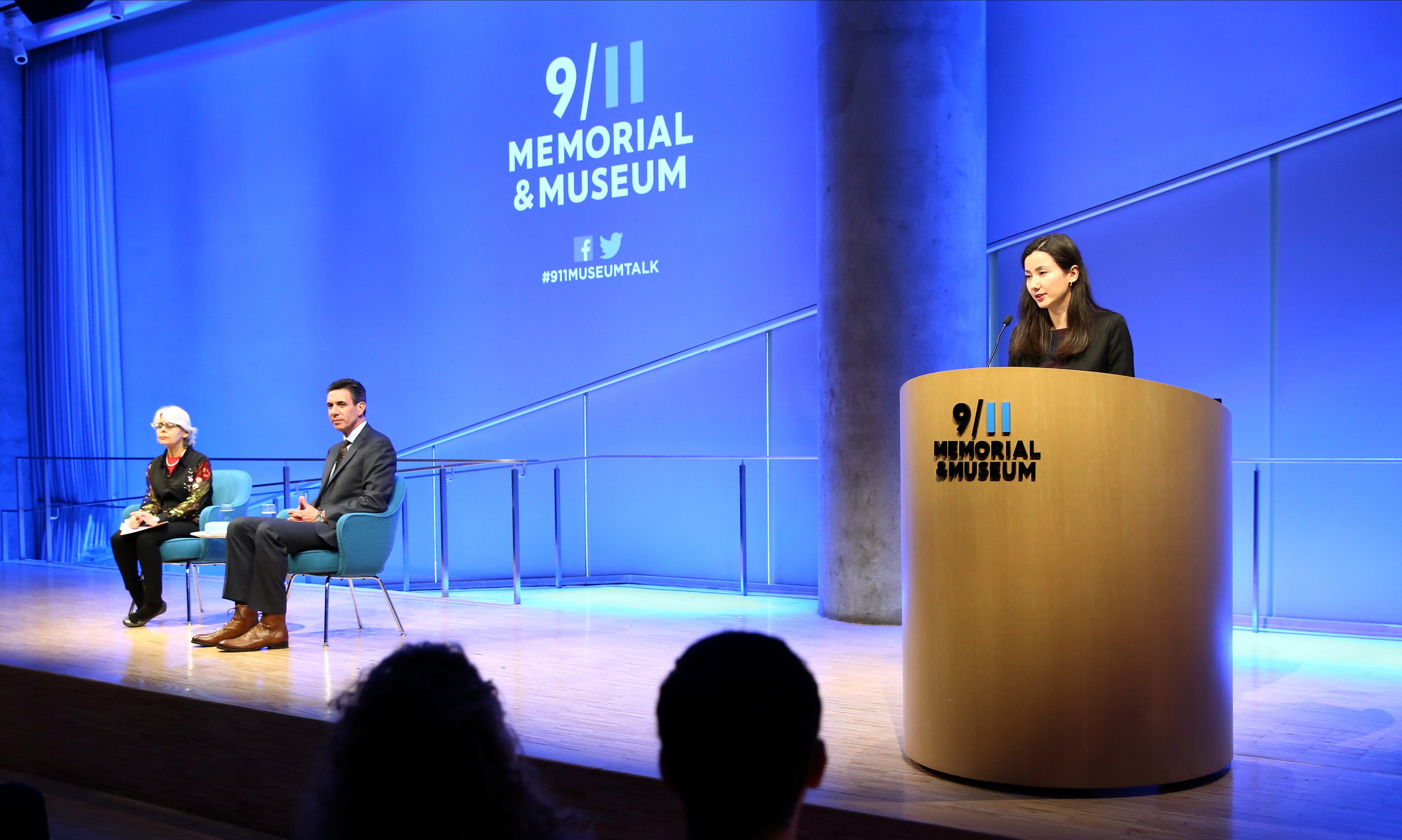 A man and a woman sit on stage during a public program in the Museum auditorium. A second woman stands at a wooden podium, speaking to the audience. Blue light fills the wall behind the stage and a projection on the wall reads “9/11 Memorial & Museum.”