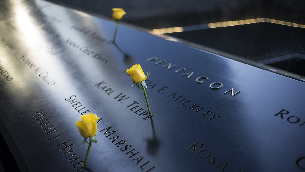 Three yellow roses stand at the names of victims etched on a sunlit Memorial. The names are near the inscription indicating Pentagon victims. The water of a reflecting pool can be seen further afield.