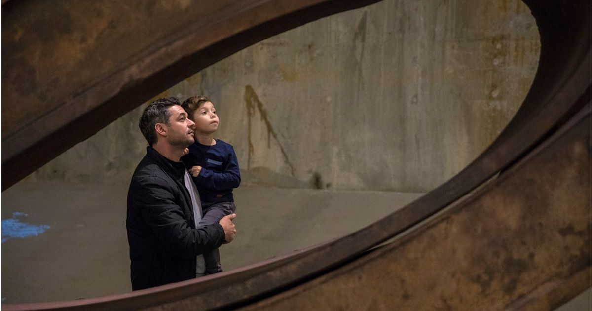 A man holds a boy as the two observe an object out of view in Foundation Hall. The two of them are framed by a piece of bent structural beam from the World Trade Center.