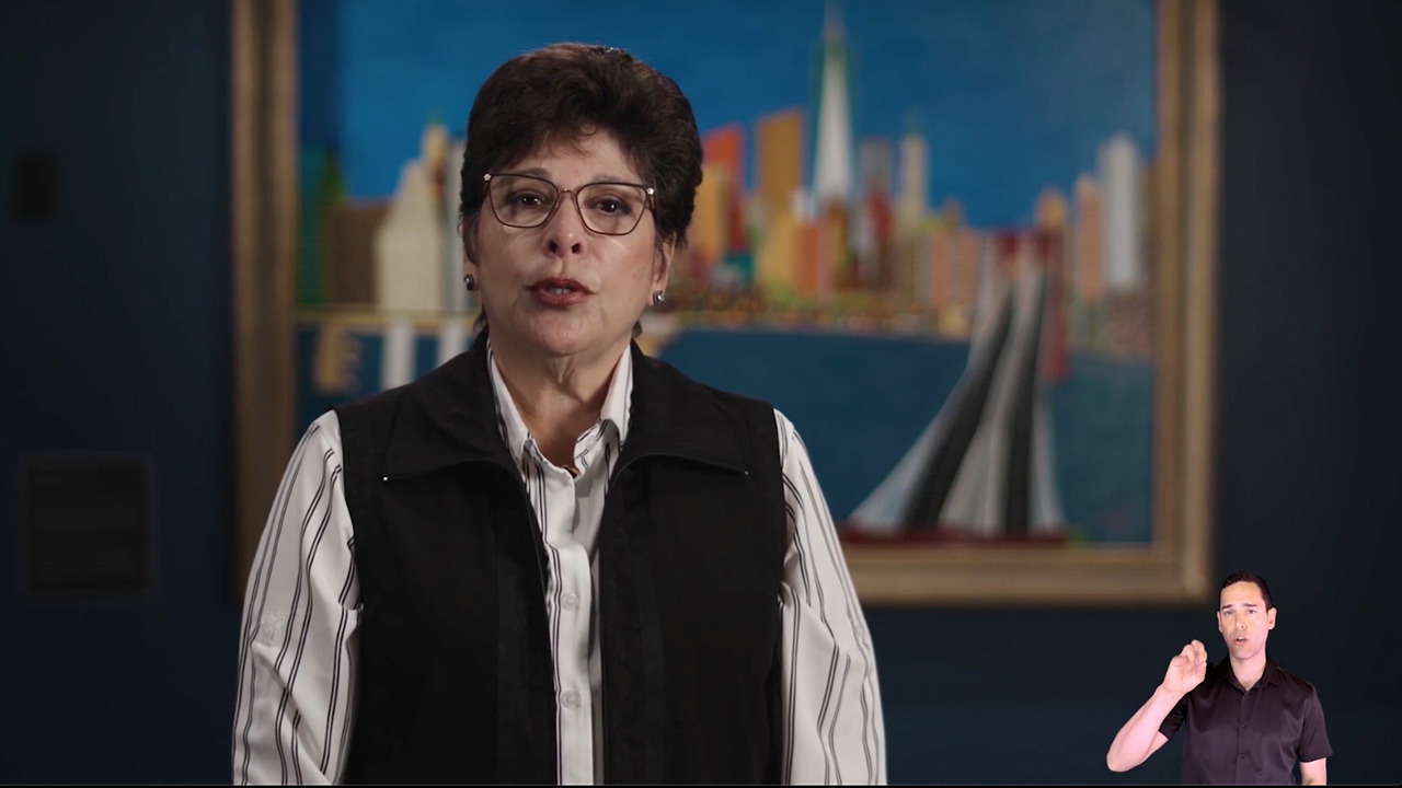 A woman with short black hair and glasses, wearing a blouse and black vest sits, talking, in front of a painting of downtown Manhattan. An ASL interpreter is shown in the lower right corner of the screen.