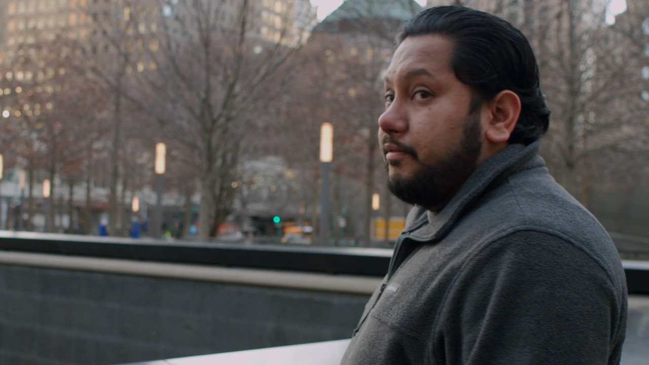 Naveed Shah has dark brown hair, brown eyes, and a mustache and beard. He stands near the 9/11 Memorial, where victims' names are engraved on a waist-high wall.