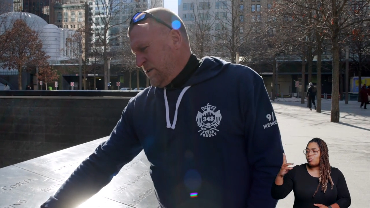 Tim Brown has short cropped hair and wears an FDNY sweatshirt. He stands at the 9/11 Memorial, where victims' names are engraved on waist-high walls. An ASL Interpreter with long hair and glasses is superimposed in the lower-right corner.