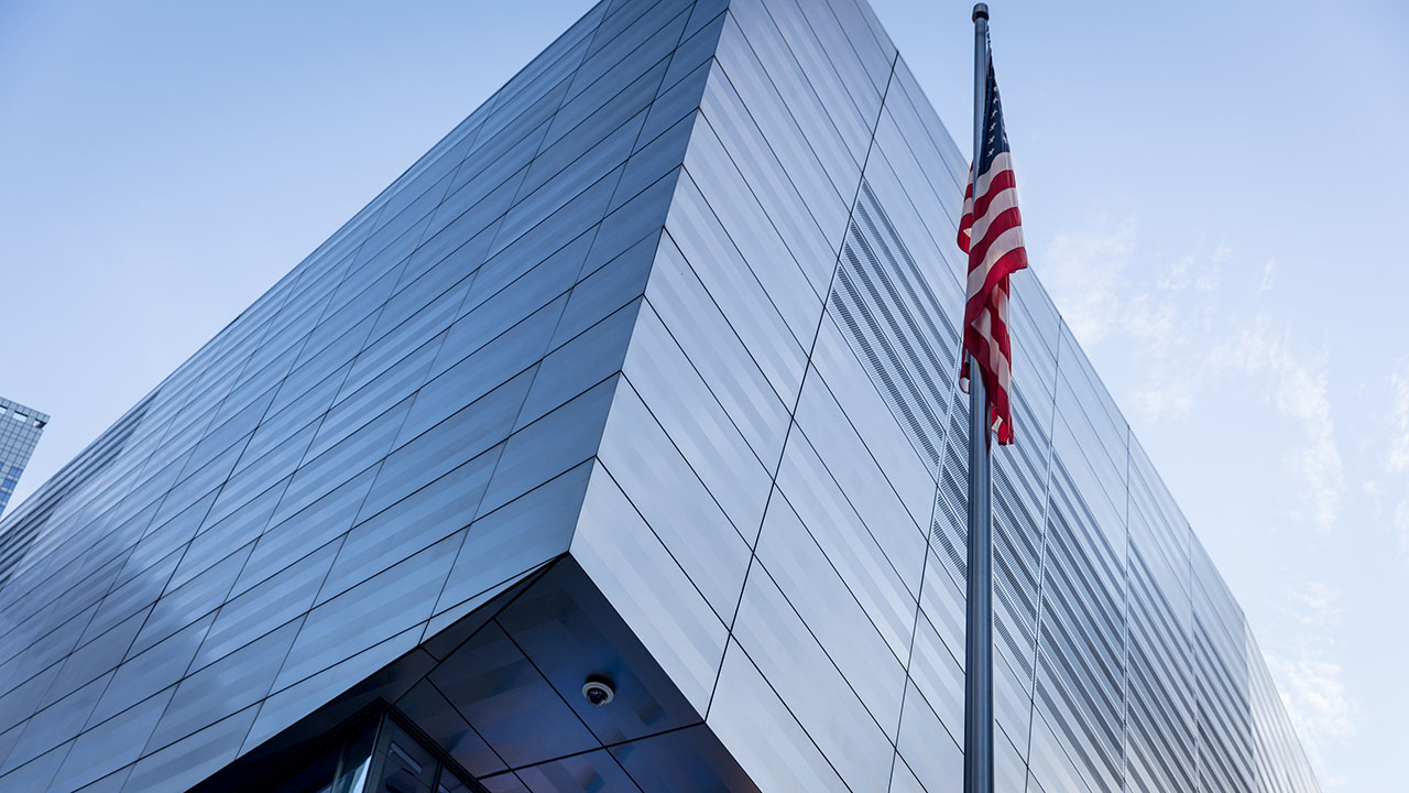A corner of the two-story, glass and steel Museum Pavilion reflects the light blue sky. An American flag is on a pole to the right.