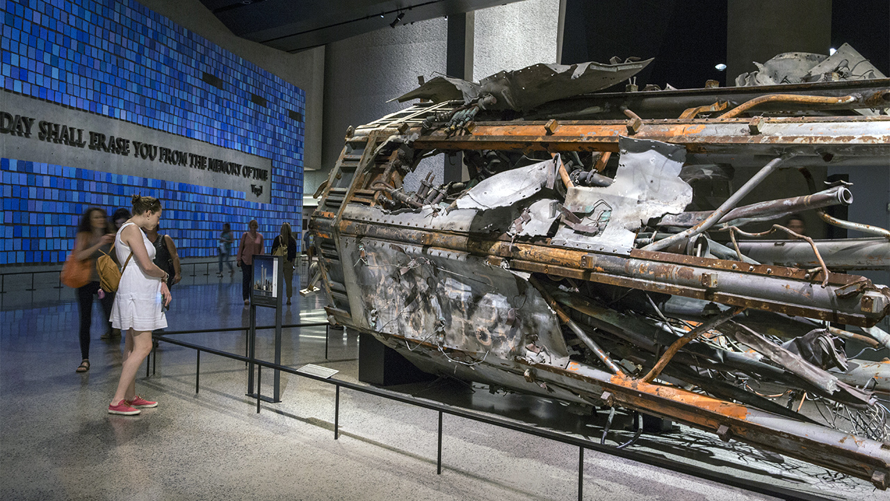 A woman in a white dress looks at a damaged portion of the North Tower’s massive broadcast antenna displayed on its side at the Museum.