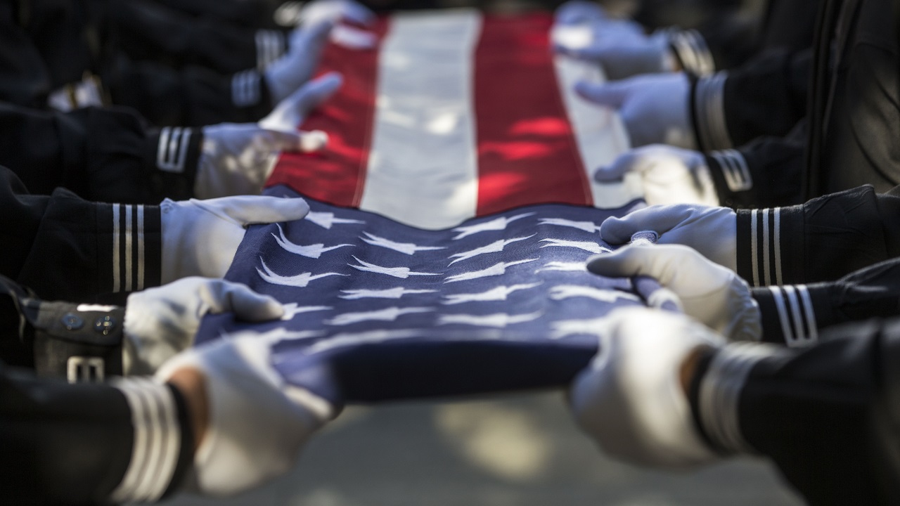 The white-gloved hands of people in black uniforms grip a folded American flag during a ceremony. Spots of sunlight and shadows fall on the flag.