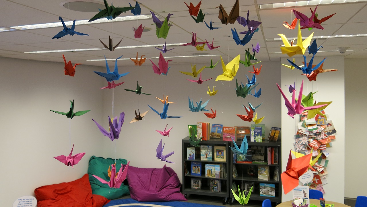Numerous paper origami cranes of many colors hang from a classroom ceiling. In the background is a bookcase and several brightly colored pillows propped against the classroom wall.