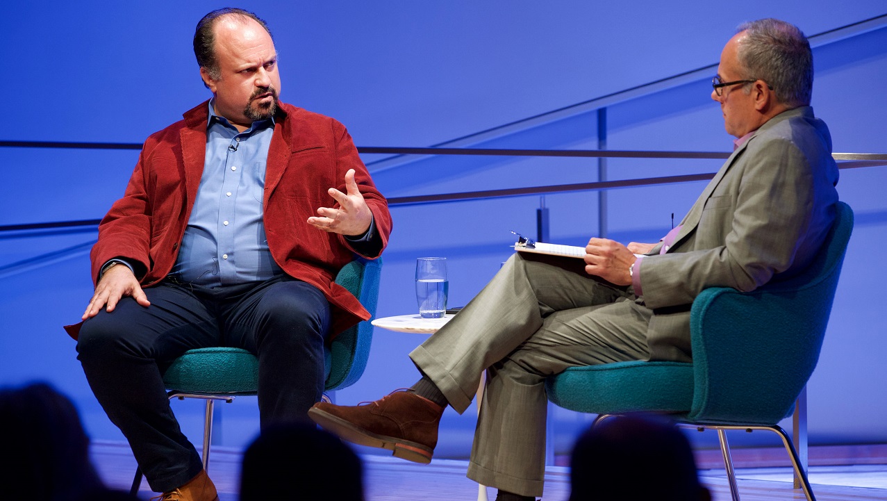 Two men speak on a blue-lit auditorium stage. The heads of the audience members appear in silhouette in the foreground.