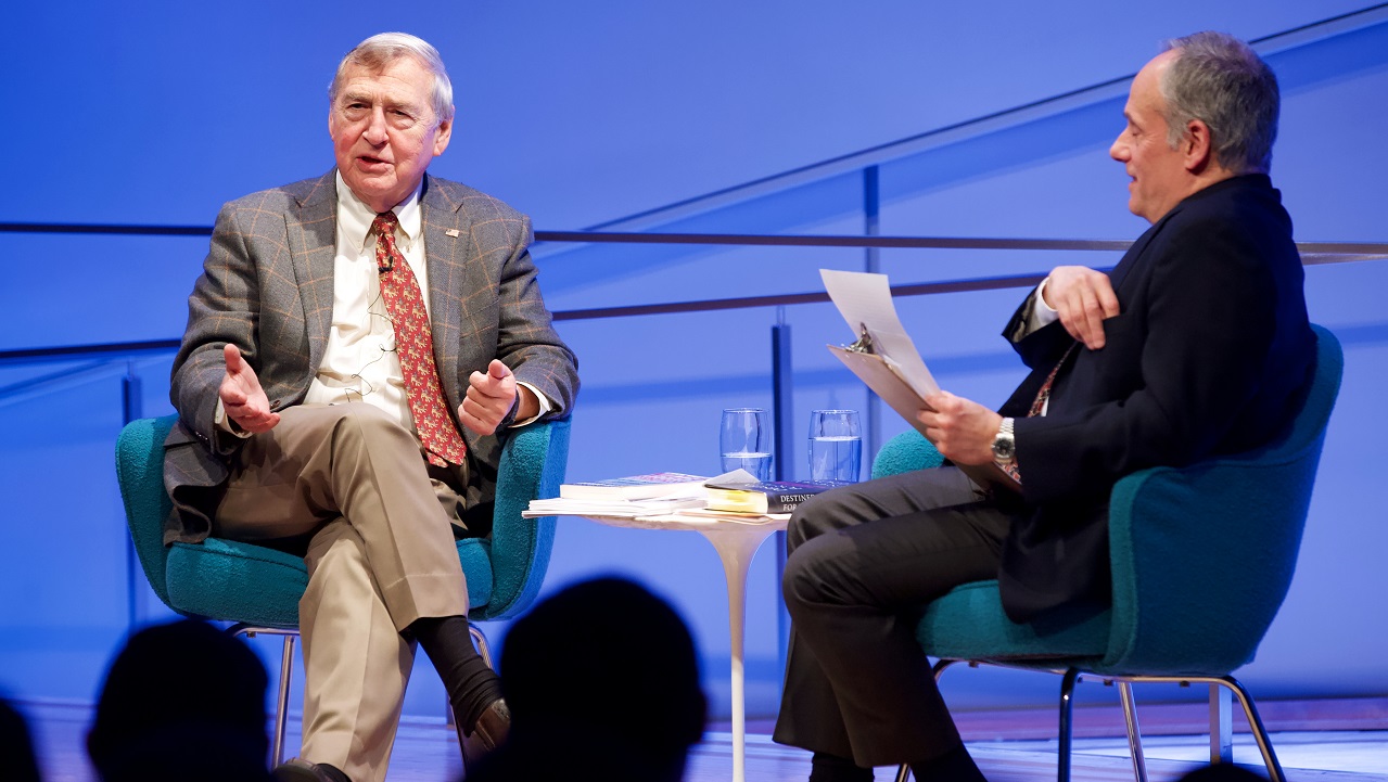 Two men sit on a blue-lit auditorium stage. One man gestures with his hands and addresses the audience, who appear in the foreground in silhouette.