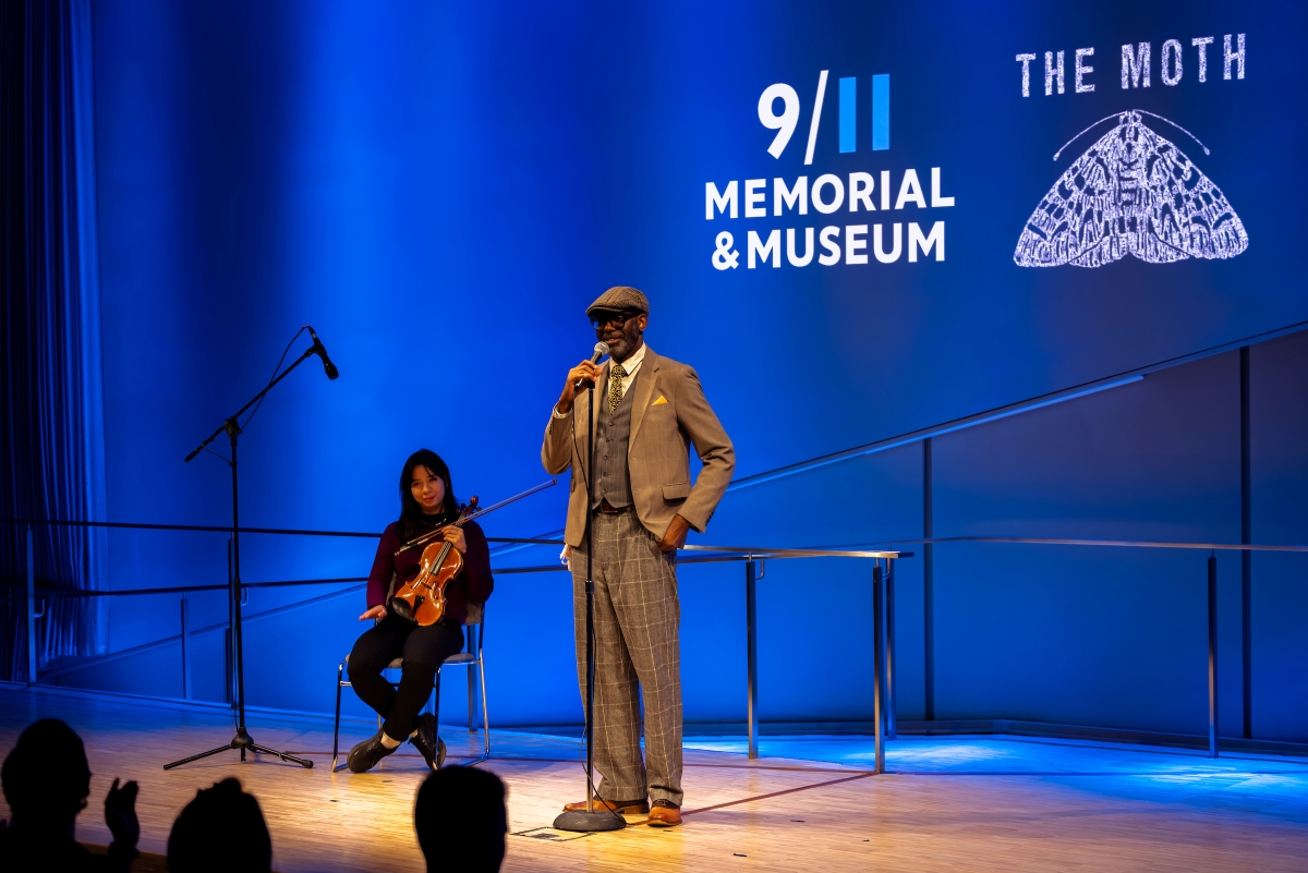 Moth Host and Violinist are on stage in front of a blue background 911 and MOTH logos