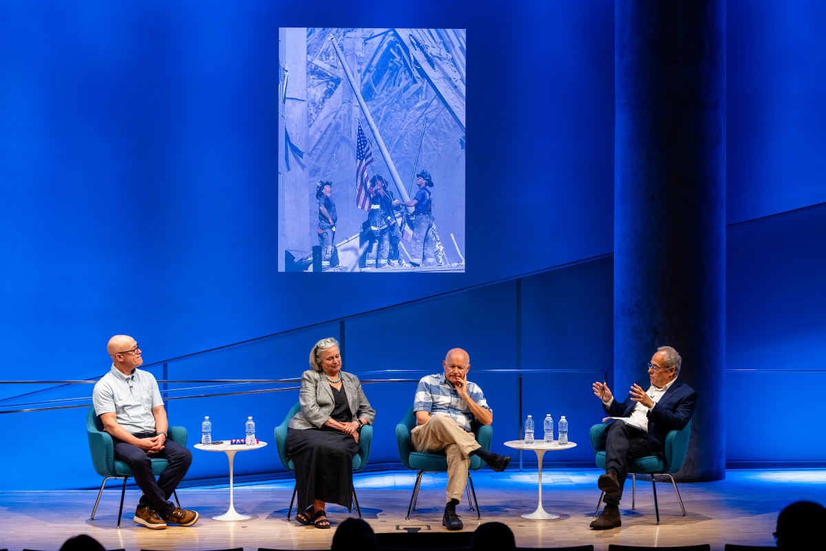four people sit on stage in front of a blue background with a large photo on the screen -- the flag from Ground Zero