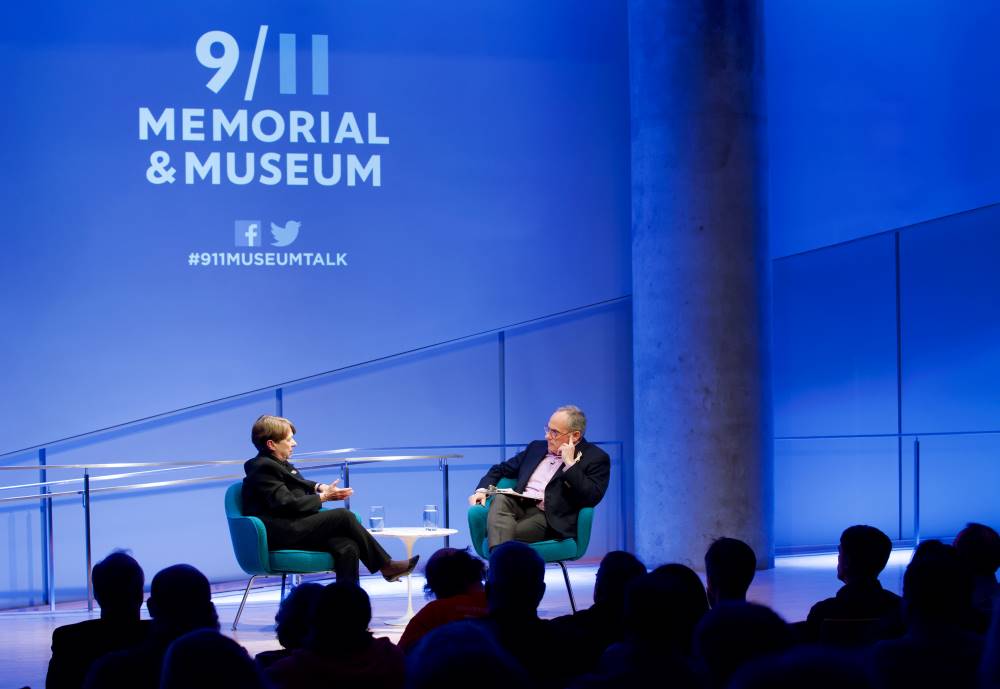 Former U.S. Attorney Mary Jo White of the Southern District of New York and Clifford Chanin, the executive vice president and deputy director for museum programs, speak onstage at the Museum Auditorium under the logo of the 9/11 Memorial & Museum, which is projected on the blue-illuminated wall behind them. The silhouettes of audience members are in the foreground.