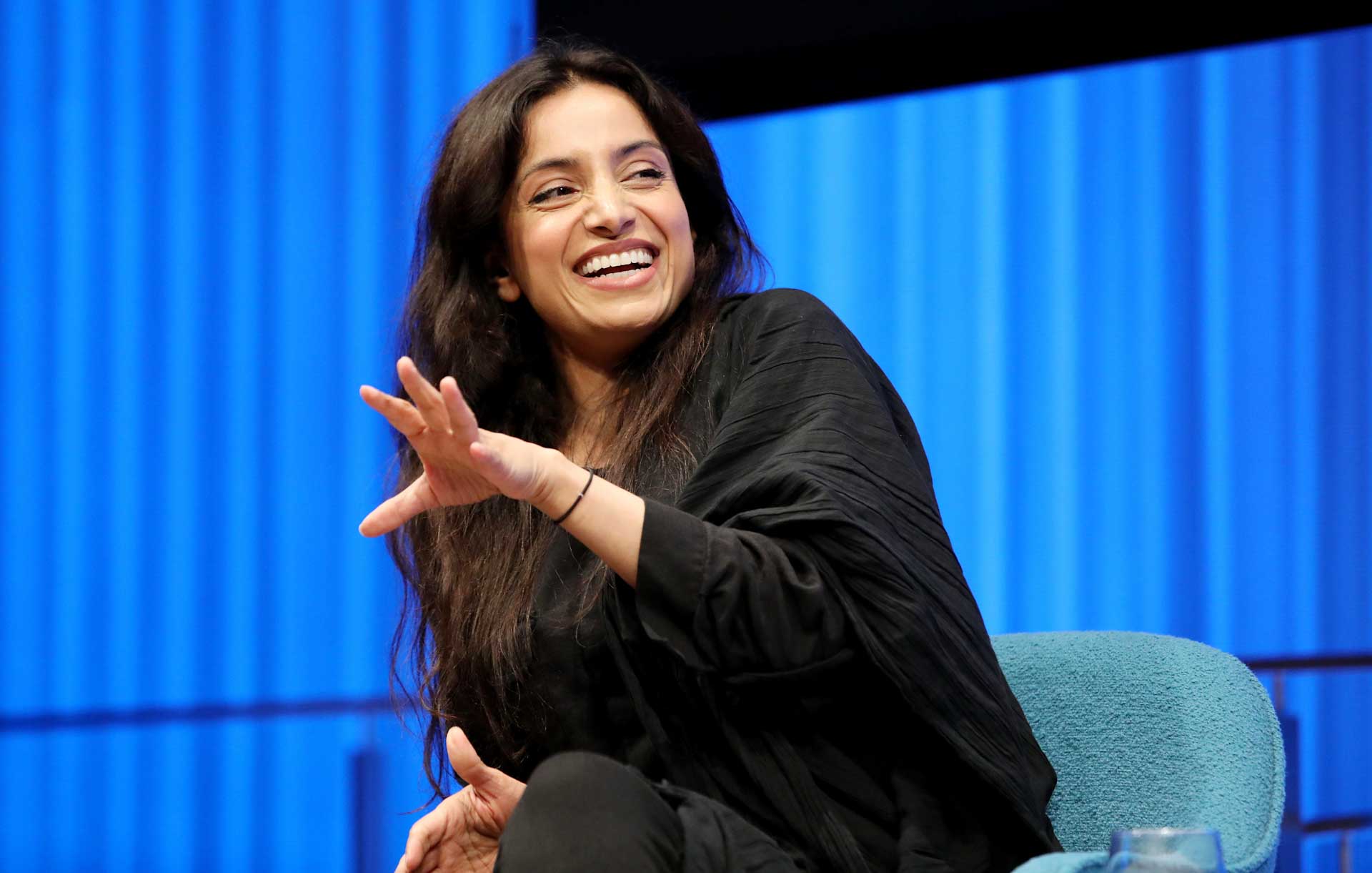 Emmy Award–winning documentarian Deeyah Khan smiles as she leans forward and gestures with her left hand onstage while speaking during a public program at the Museum Auditorium. Curtains behind her are lit up blue.