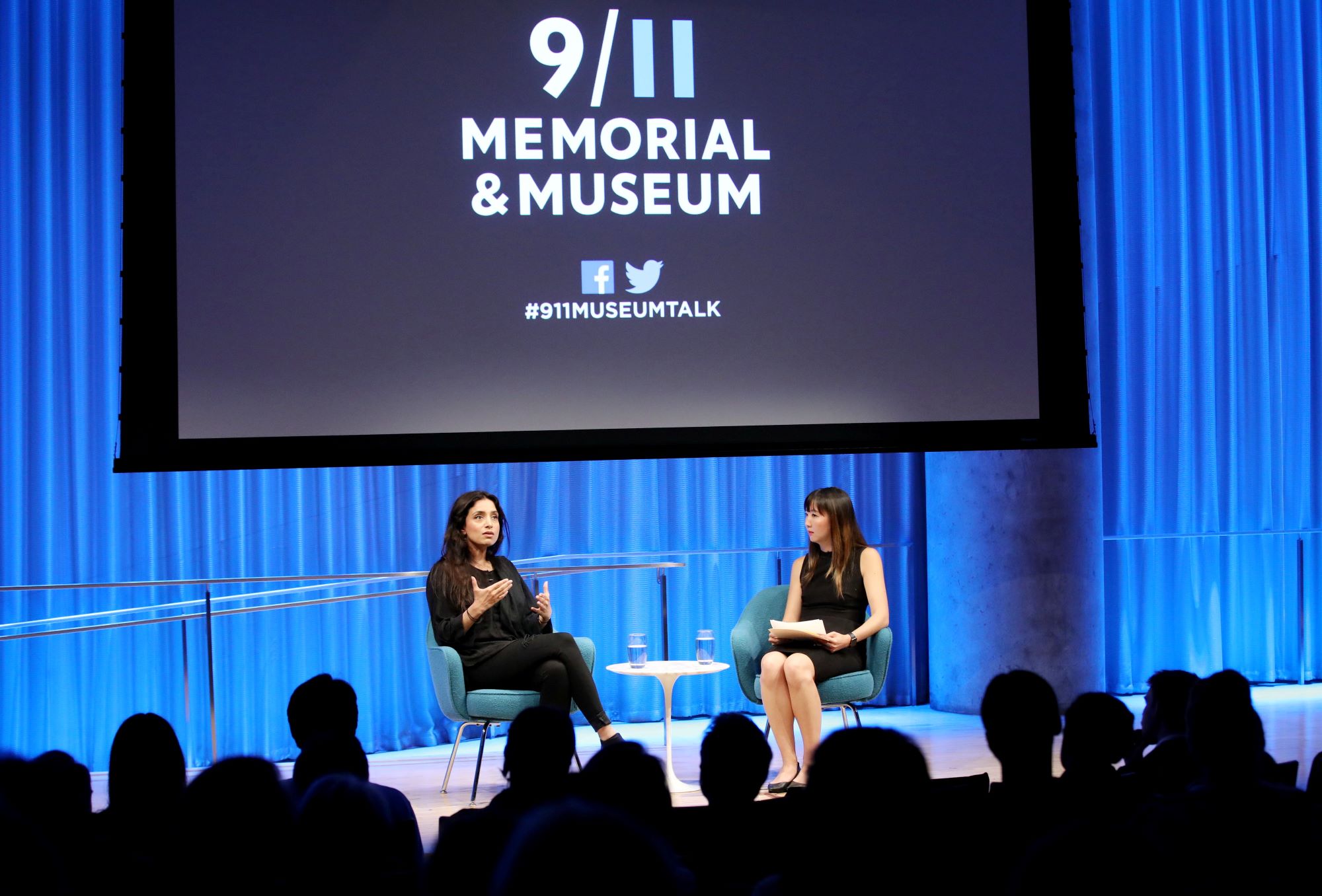 In this wide-angle photo of the Museum Auditorium stage, Emmy Award–winning documentarian Deeyah Khan speaks to the audience while seated next to a woman hosting the event. The audience members are silhouetted by blue and white lights shining on the stage. A large projector screen has been lowered above the two of them.