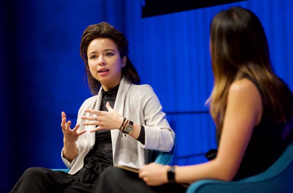 VICE correspondent Isobel Yeung gestures as she speaks onstage next to a woman hosting a public program at the Museum Auditorium. A curtain behind her is lit up blue.