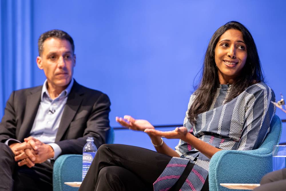 Vidhya Ramalingam, founder of Moonshot CVE, holds out her hands as she speaks onstage at the Museum Auditorium. Hany Farid, Dartmouth computer science professor and senior advisor to the Counter Extremism Project, listens with his hands in his lap in the background.