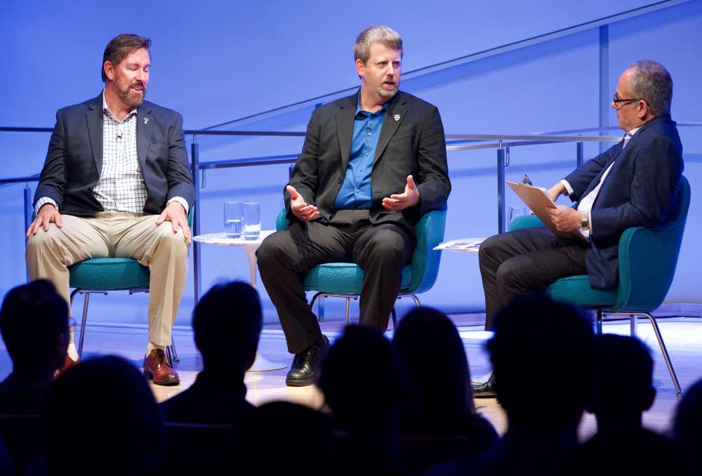 Former special forces commander Maj. Mark Nutsch speaks onstage at the Museum Auditorium as he sits next to former special forces commander retired Master Sgt. Scott Neil. Clifford Chanin, the executive vice president and deputy director for museum programs, sits to their left holding a clipboard and listening on. Audience members are silhouetted in the foreground.