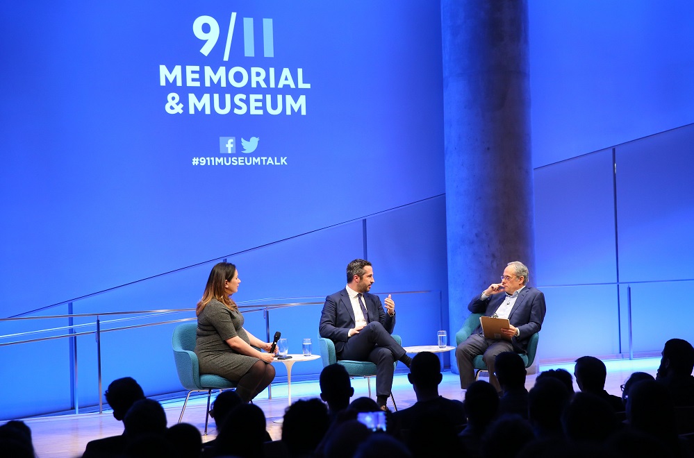 Lorenzo Vidino, director of the George Washington University Program on Extremism, speaks onstage at the Museum Auditorium while seated next to New York Times foreign correspondent Rukmini Callimachi and Clifford Chanin, the executive vice president and deputy director for museum programs. Vidino faces Chanin as he speaks and gestures with his left hand. The wide-angle photo includes the audience, who are silhouetted by bright white and blue lights from the stage.