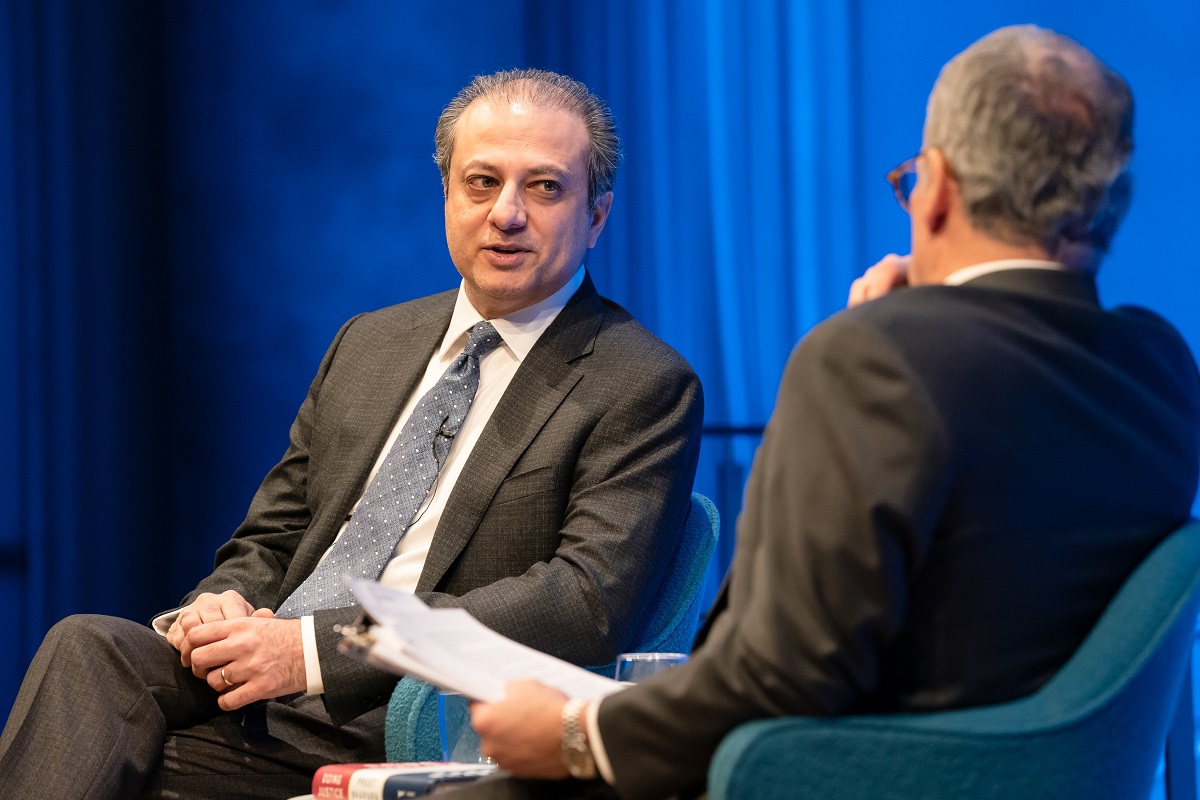 In this close-up view of the Museum Auditorium stage, Preet Bharara, the former U.S. attorney for the Southern District of New York, speaks with moderator Clifford Chanin, who is out of focus and has his back turned to the camera. A wall behind Bharara is lit blue from the stage lights.