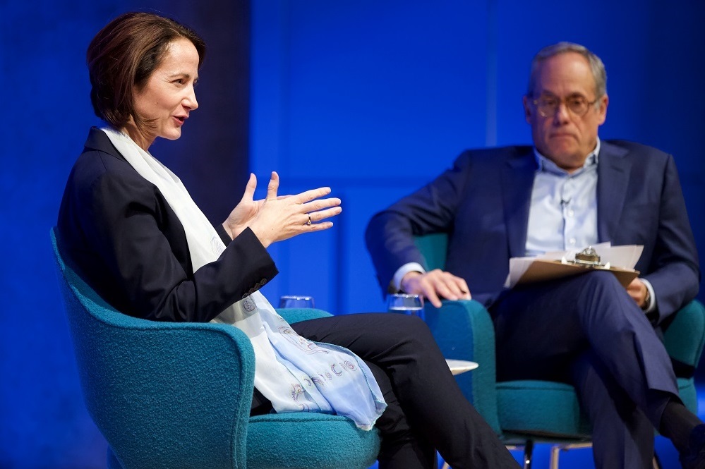 A woman wearing a gauzy scarf and a navy-blue suit appears in profile on a blue-lit auditorium stage. She gestures with her hands and looks ahead to address an unseen audience. A man, moderating with a clipboard in hand, stares off into the distance.