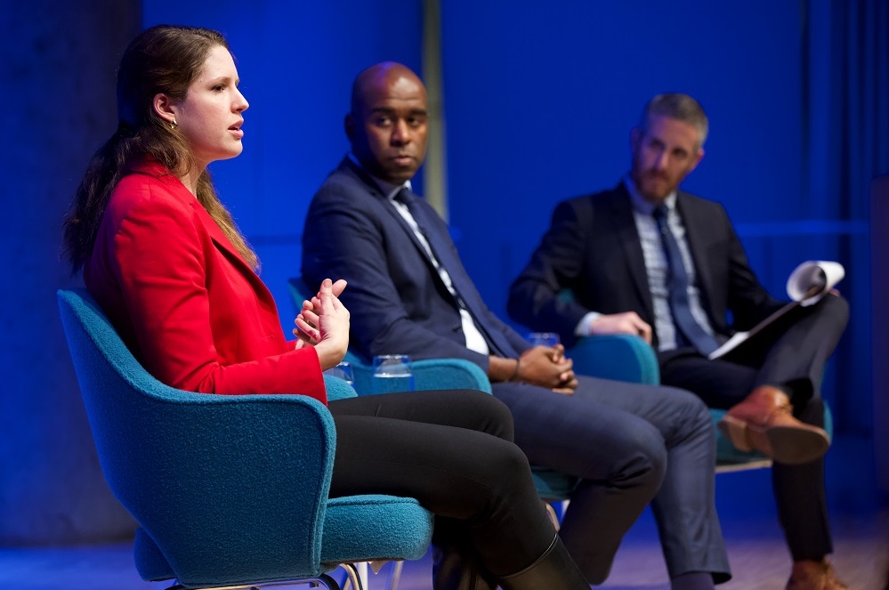 In this close-cropped photograph, three public program participants sit on a blue-lit auditorium stage. Closest to the camera sits a woman in a red blazer, who appears in profile, and addresses an unseen audience. The other two men on stage watch her and listen.