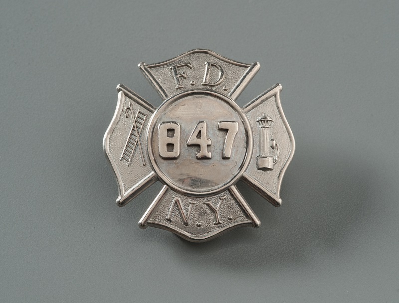 FDNY badge shaped as a Maltese cross with the numbers '847' in the center. The letters F D N Y and images of a hook, ladder, and fire hydrant fill the four cross arms.