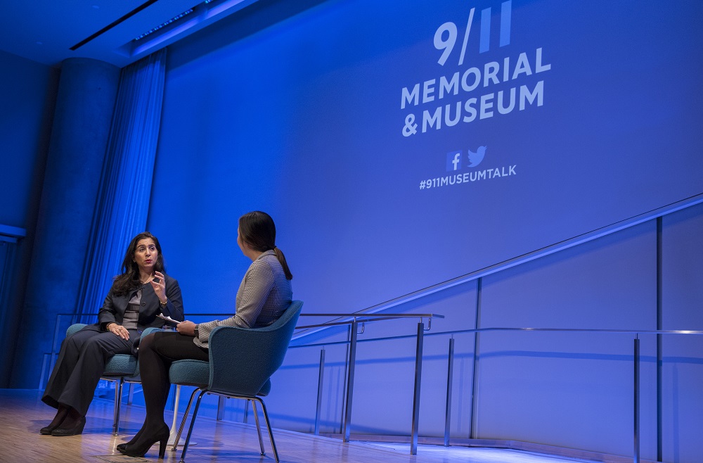 Two woman sitting on a blue-lit auditorium stage around photographed from the side of the stage. One woman in a dark gray suit gestures with her hands while another woman in a light gray blazer listens with a clipboard in her lap.