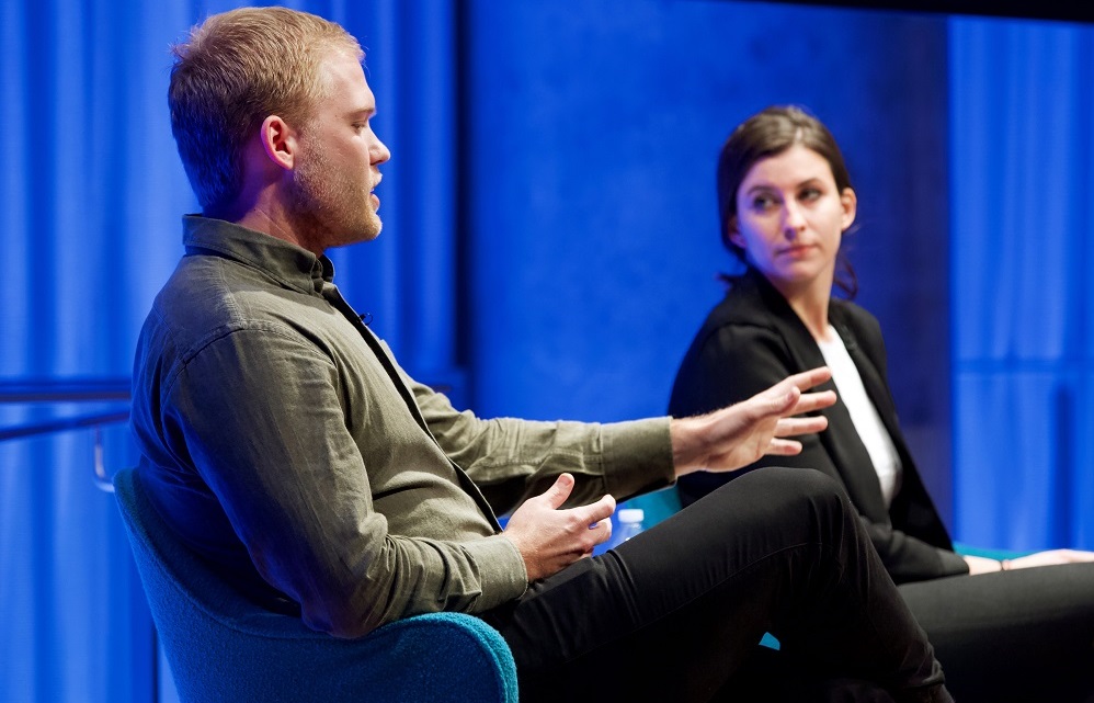 A man on a blue-lit auditorium stage speaks while a woman to his left looks on.