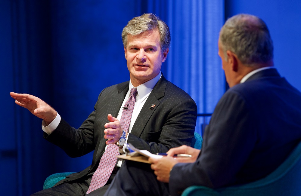 Two men in suits sit on a blue-lit auditorium stage. One gestures with his hands while the other looks on.