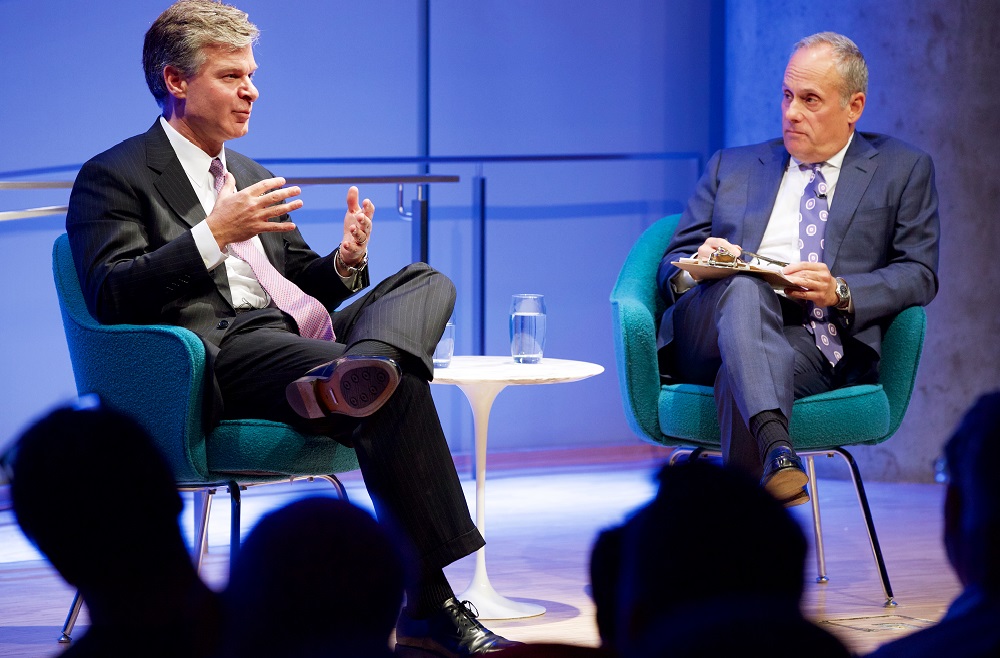 Two men sit with crossed legs on a blue-lit auditorium stage. Heads of audience members appear in silhouette in the foreground.