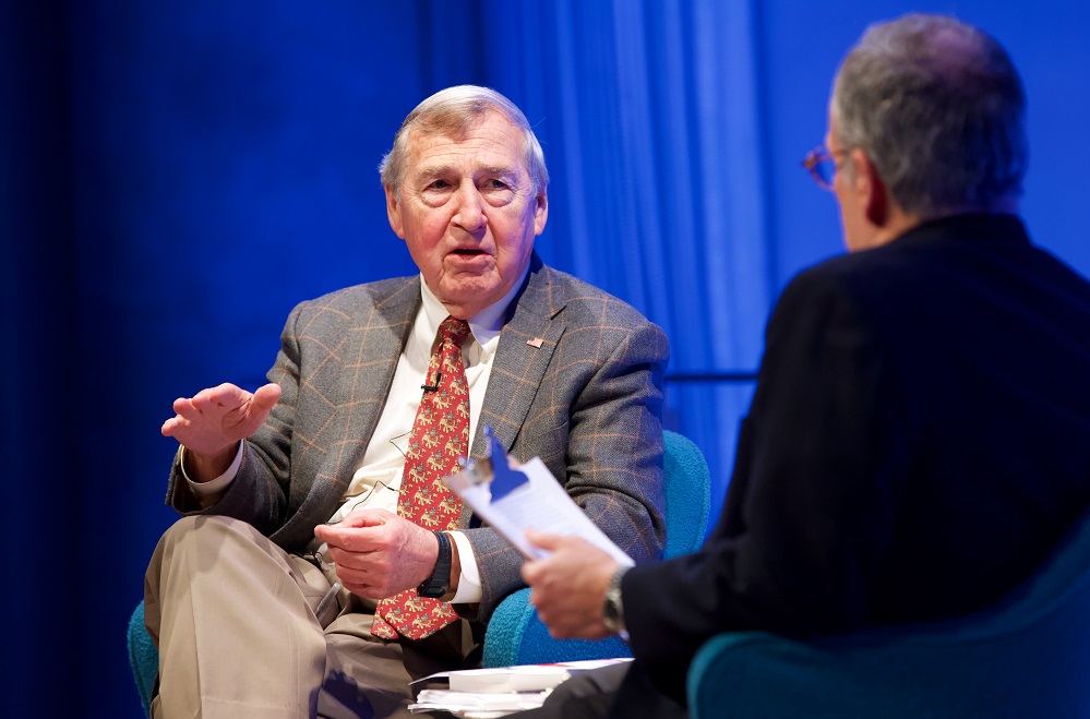 Two men sit on a blue-lit auditorium stage. One man gestures with his hands and addresses the other.